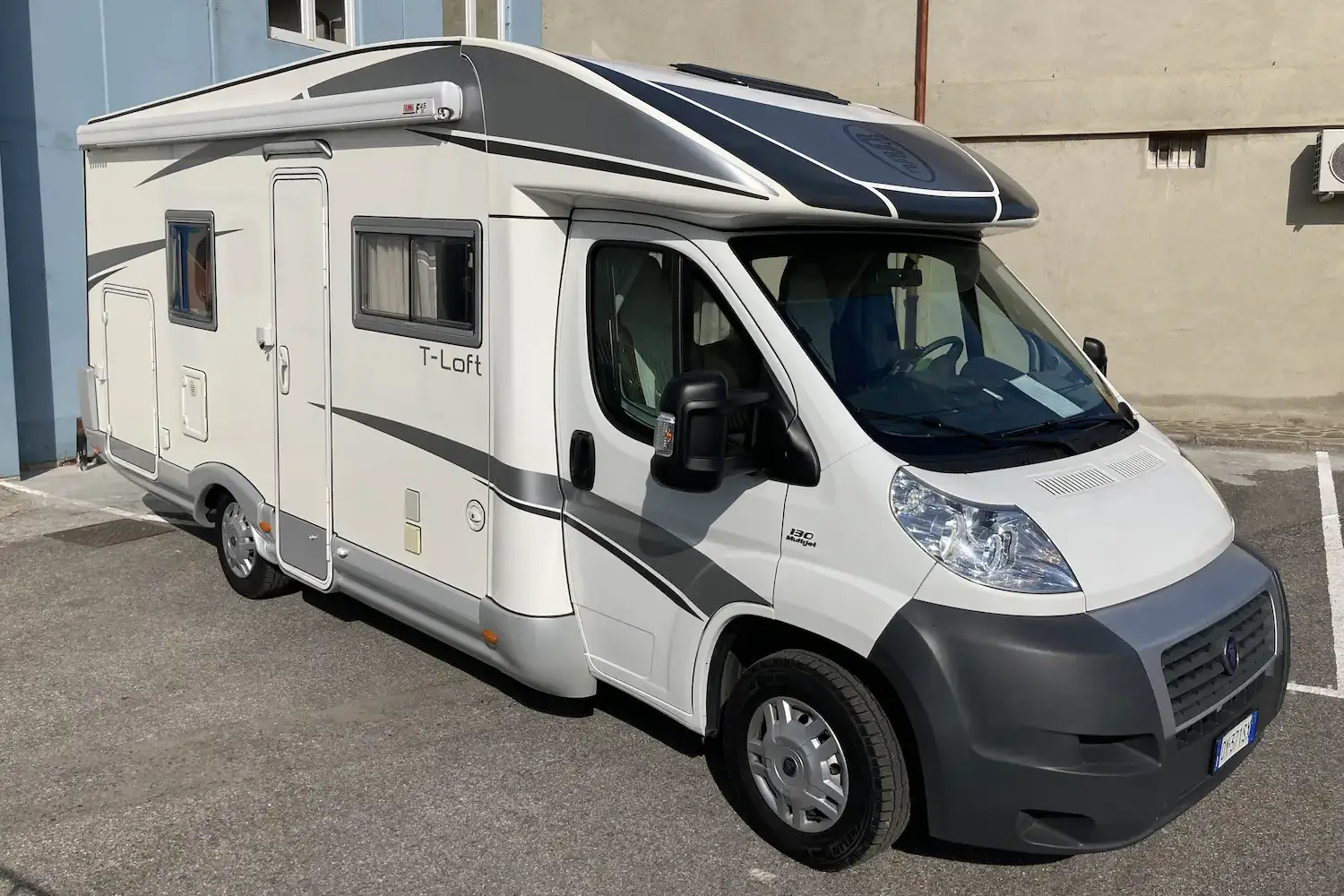 Camper mansardato modello T-Loft su base Fiat Ducato, carrozzeria bianca con fasce grafiche grigie e tetto rialzato, parcheggiato in un piazzale.