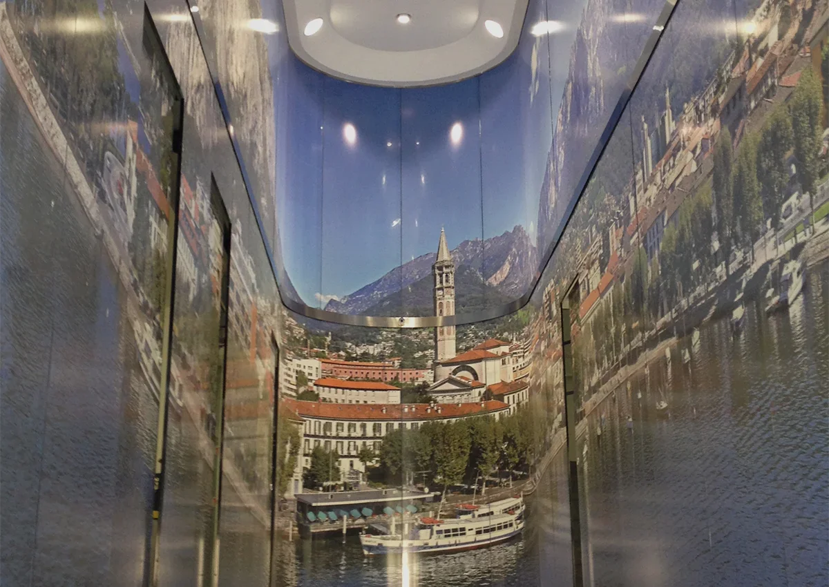 Corridoio interno con pareti rivestite da una stampa fotografica panoramica su vetro raffigurante il lungolago di Lecco con il centro storico, le montagne sullo sfondo e le imbarcazioni ormeggiate.