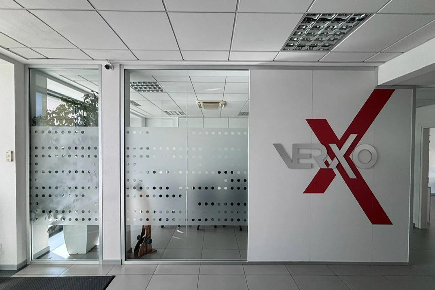 Ingresso di ufficio con parete bianca che riporta il logo Verxo in rosso e grigio, affiancata da pareti divisorie in vetro satinato con motivo a pois e pavimento in piastrelle grigie.