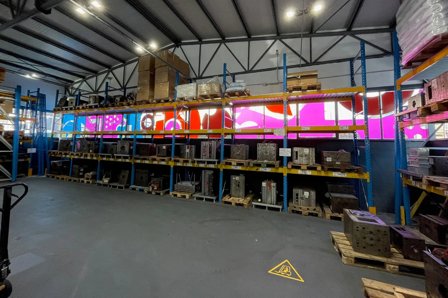 Interno di un magazzino industriale con scaffalature metalliche blu e gialle cariche di stampi e attrezzature su pallet, finestre rivestite con pellicola colorata a motivi grafici fucsia, rosso e blu.