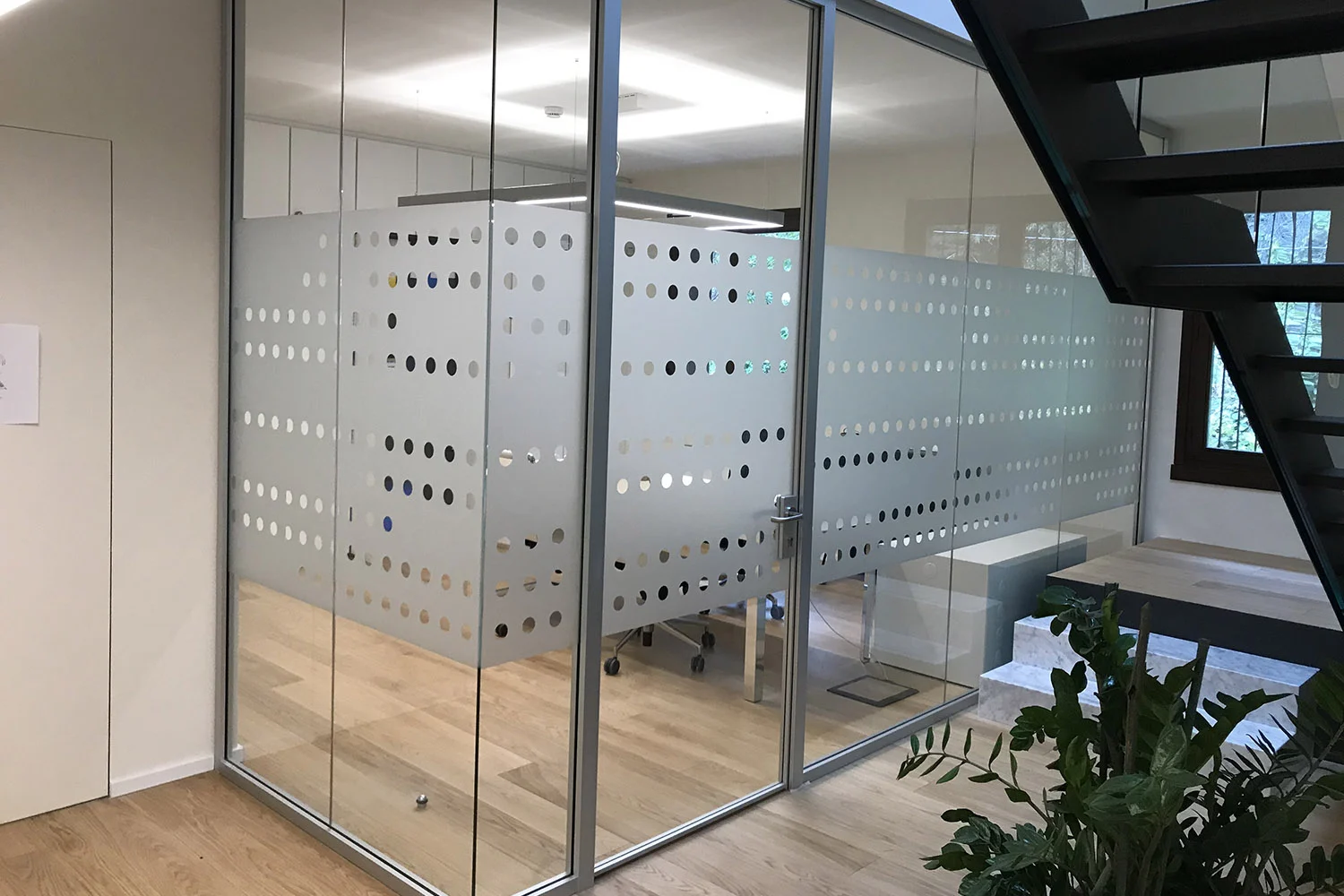 Ufficio con pareti divisorie in vetro satinato decorate con motivo a pois, pavimento in parquet chiaro, pianta decorativa in primo piano e scala in metallo sul lato destro.