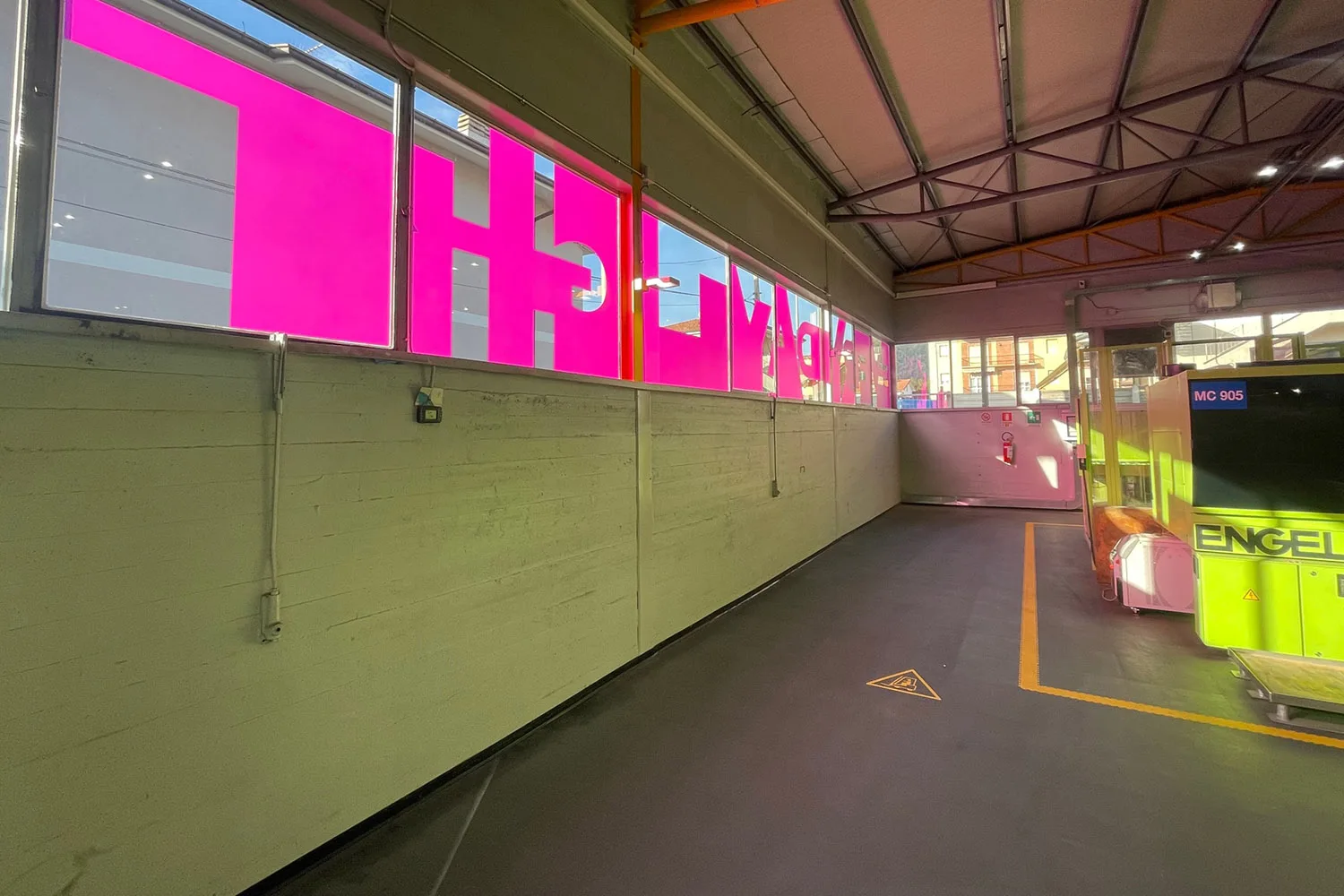 Interno di un capannone industriale con finestre rivestite da pellicola fucsia con grafica tipografica a grandi lettere, pavimento in cemento e macchinario industriale giallo sul lato destro.