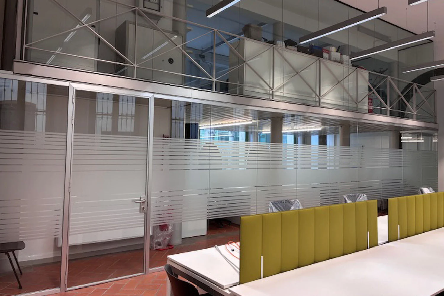 Spazio di lavoro in un edificio industriale ristrutturato con pareti divisorie in vetro satinato a strisce orizzontali, pannelli fonoassorbenti gialli, soppalco metallico e pavimento in cotto.