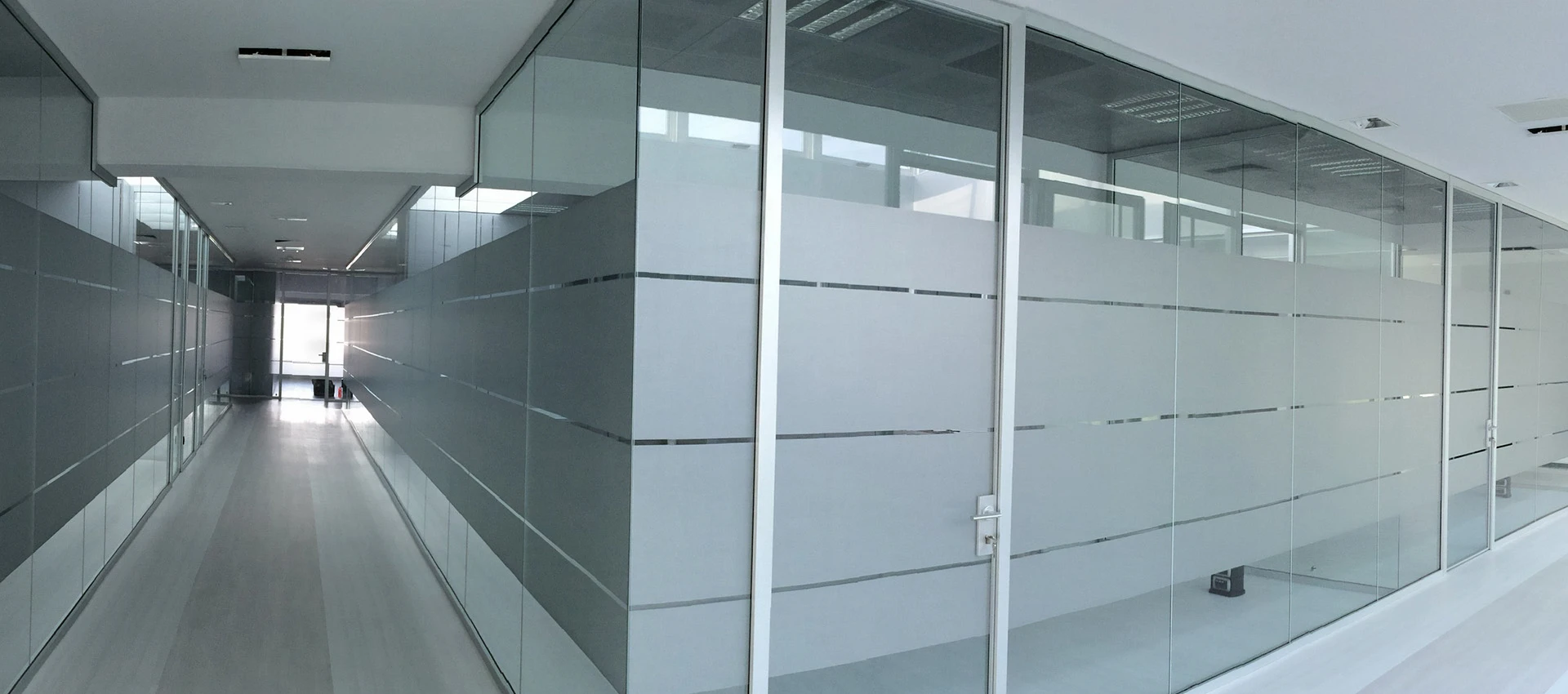 Corridoio interno di un edificio moderno con pareti divisorie in vetro satinato e alluminio, illuminazione a soffitto e pavimento in parquet chiaro.