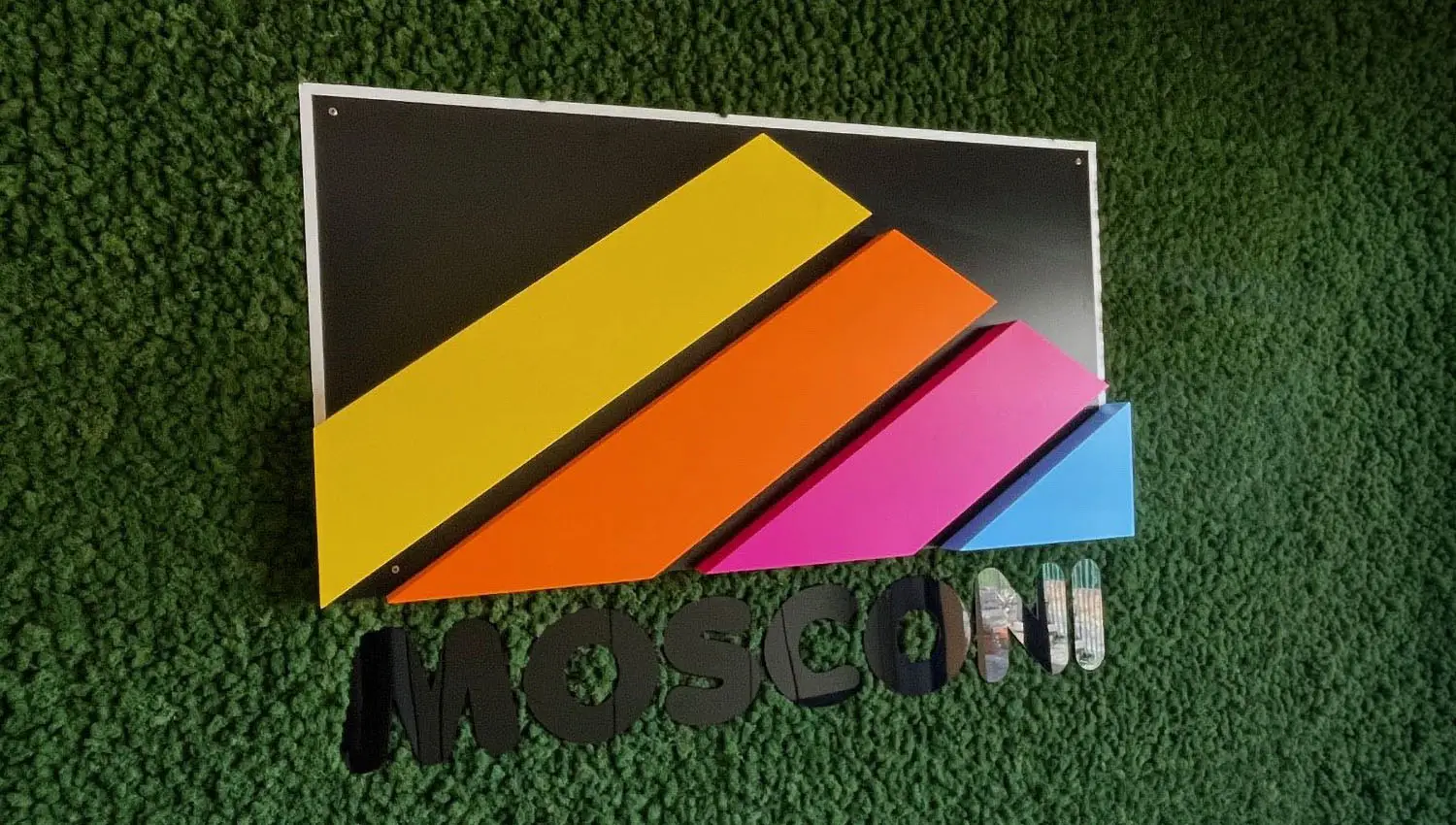 Insegna Mosconi con pannello grafico multicolore in giallo, arancione, rosa e azzurro su parete in erba sintetica verde, con lettere tridimensionali nere.